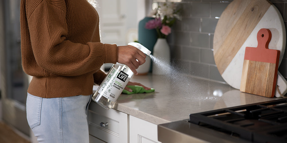 A woman spraying a bottle of Better Life cleaner on a kitchen counter.