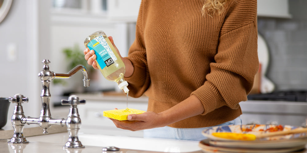 A woman is washing dishes with a Better Life product in a kitchen sink.